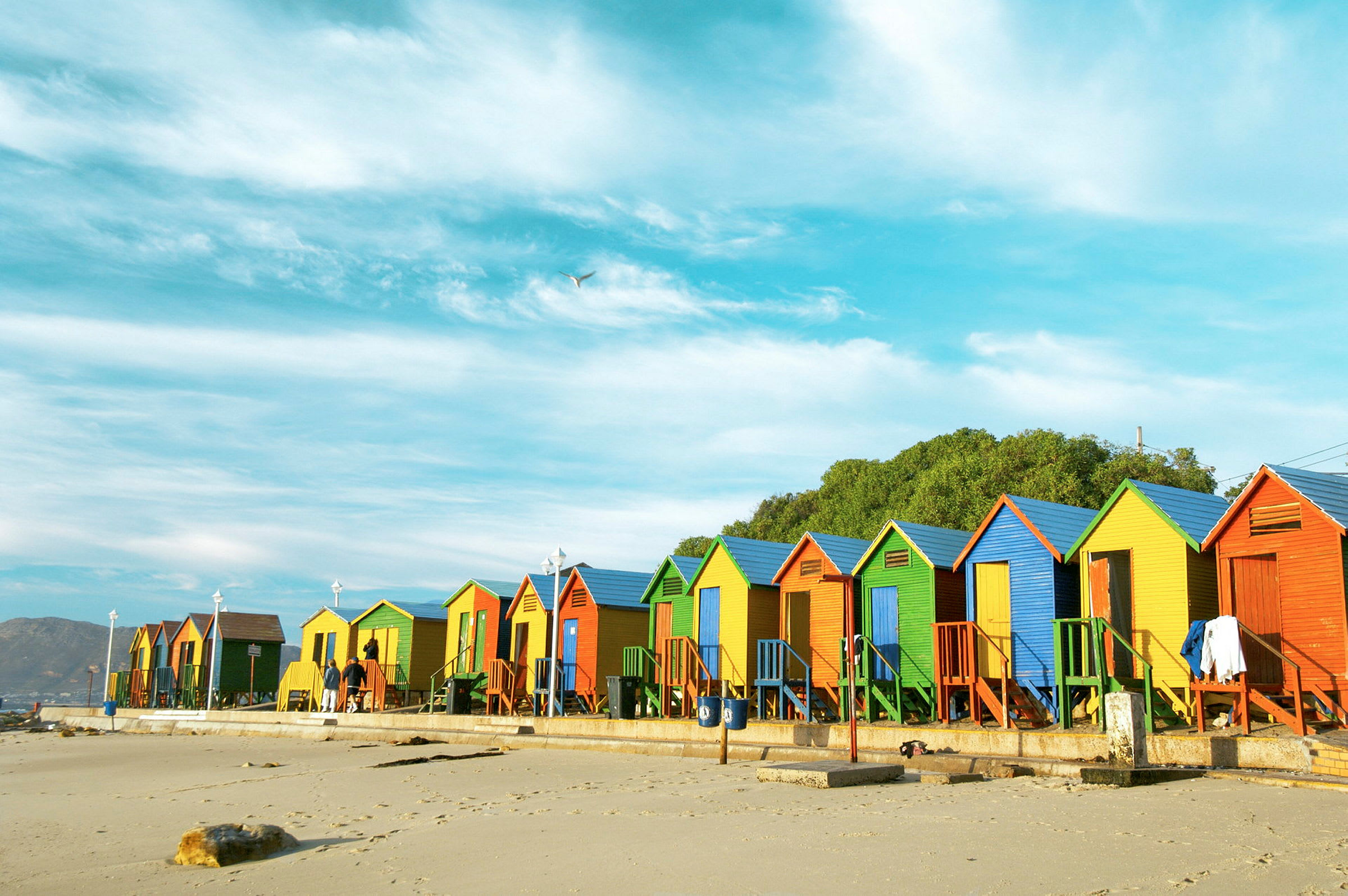 Flere fargerike strandhytter står på rekke langs en sandstrand under en blå himmel. (AI-generert tekst)