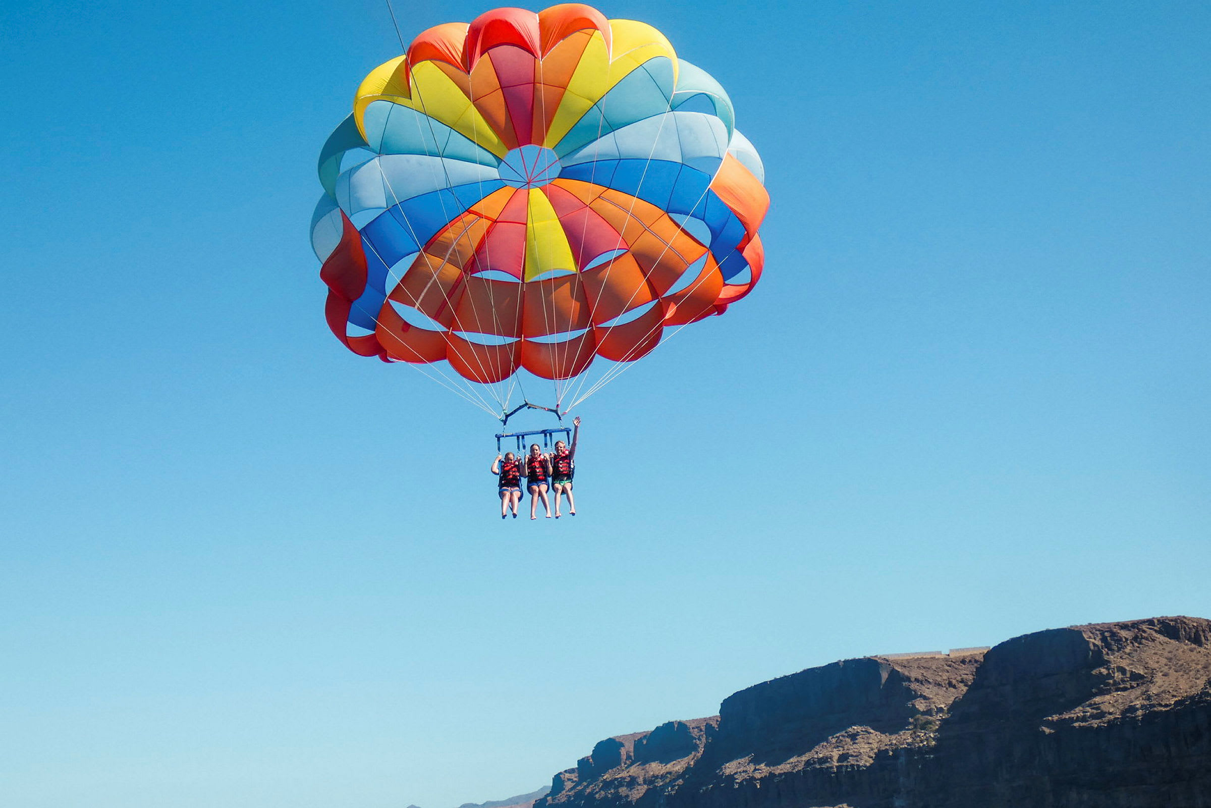 Tre personer som driver med parasailing med en stor, fargerik fallskjerm mot en blå himmel. (AI-generert tekst)