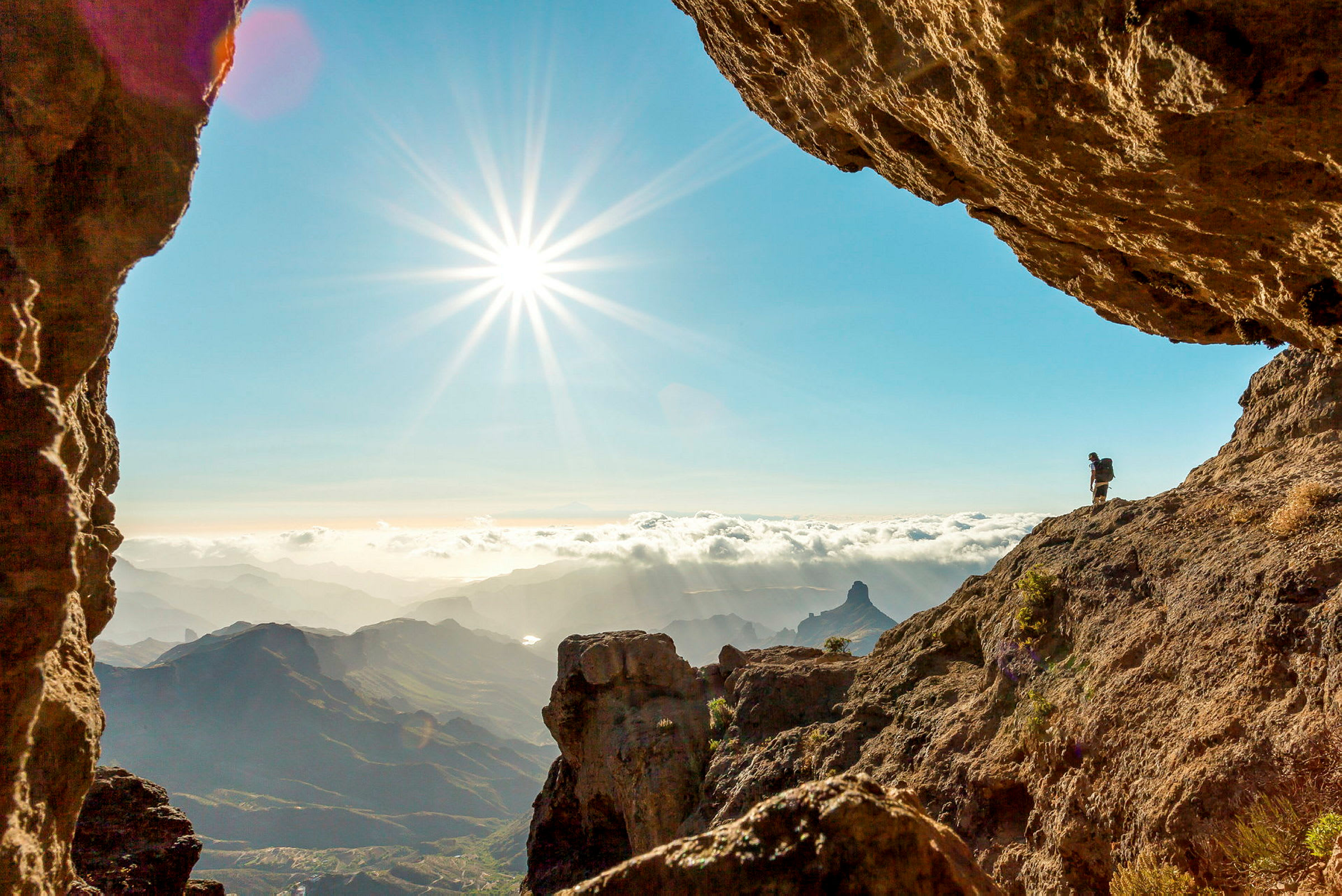 Et klippete fjellandskap med en fjellklatrer i bakgrunnen til venstre under en klar himmel med solen. (AI-generert tekst)