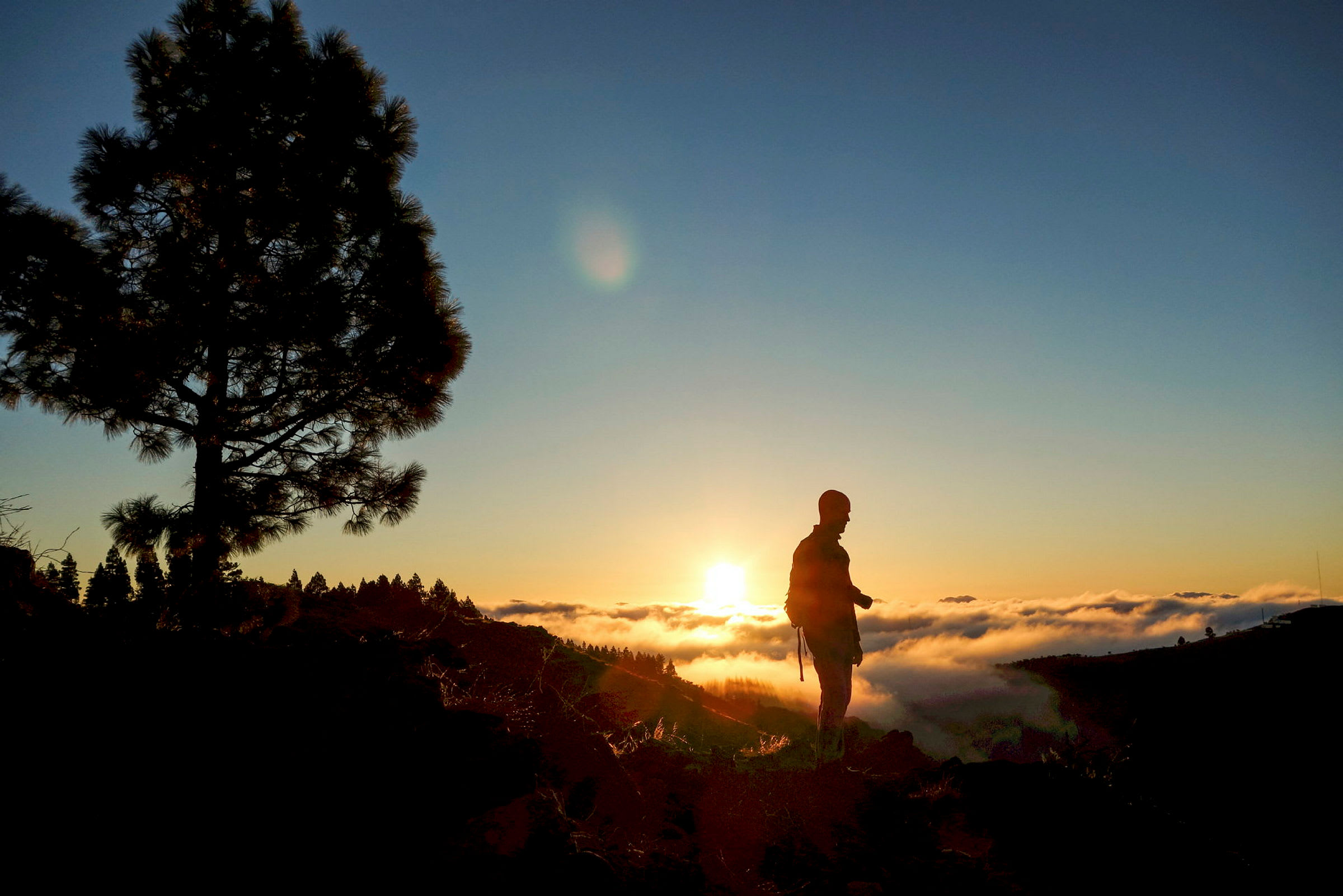 En person står på en høyde ved solnedgang med skyer i dalen nedenfor. (AI-generert tekst)