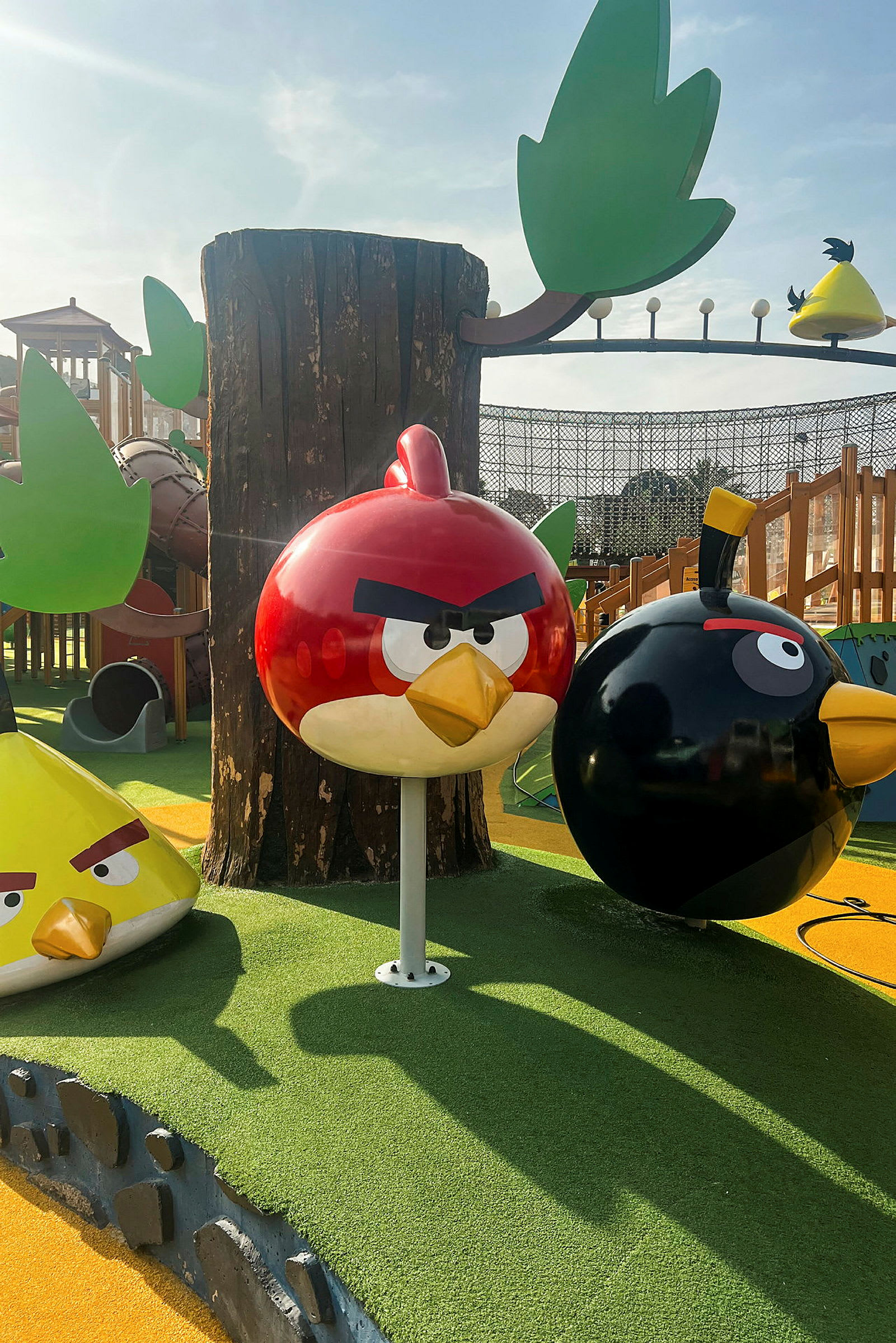 Lekeplass med Angry Birds-figurer, inkludert en rød fugl, en svart fugl og en gul trekantet fugl, med trær og andre lekestrukturer rundt. (AI-generert tekst)