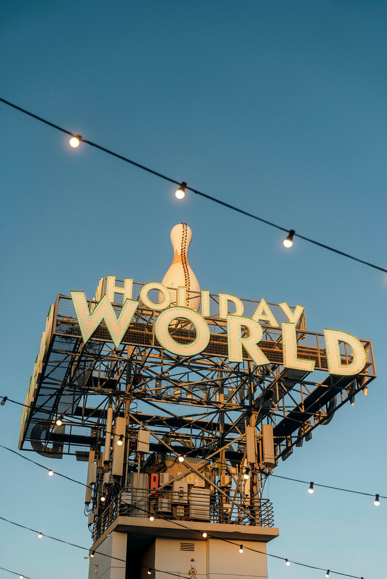 Skilt med teksten 'Holiday World' og en stor dekorert bowlingkjegle på toppen, med lyslenker mot en klar blå himmel. (AI-generert tekst)