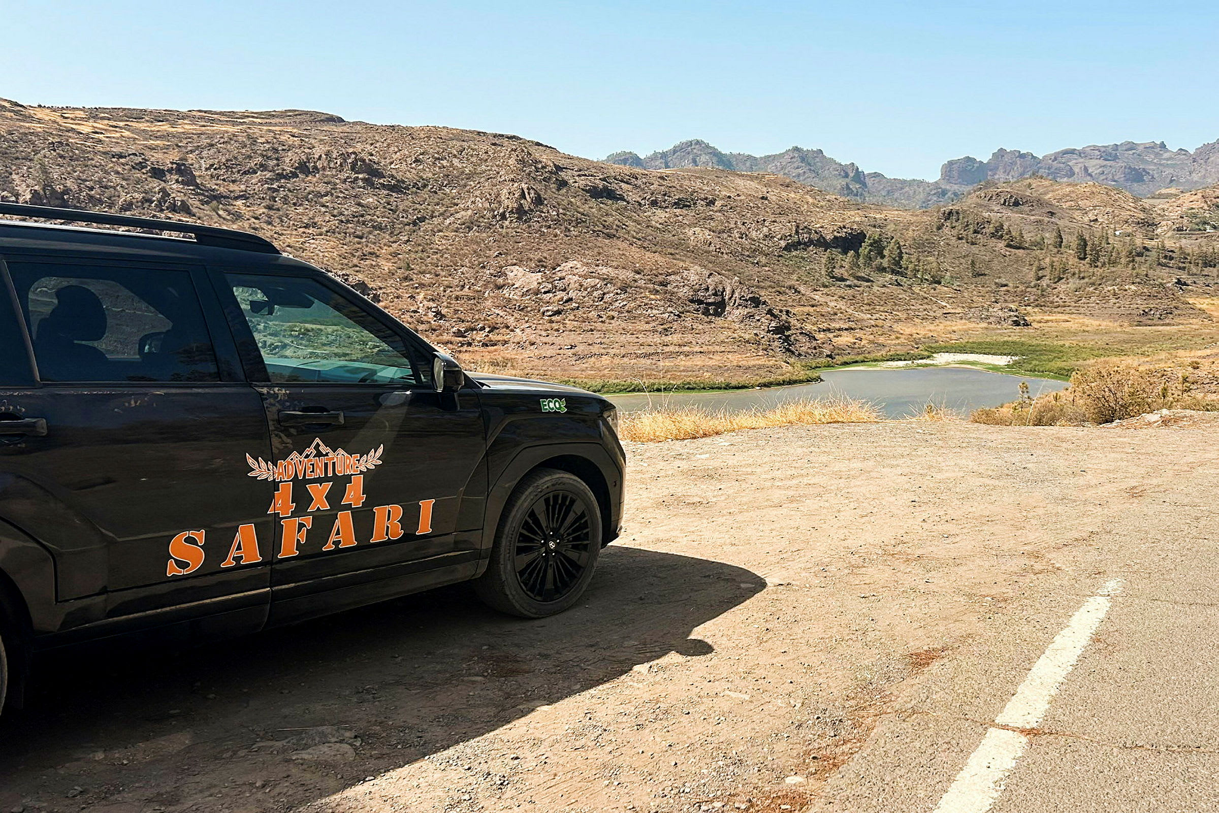 En firehjulsdrevet bil med teksten 'Adventure 4x4 Safari' parkert i et ørkenlignende landskap med en innsjø i bakgrunnen. (AI-generert tekst)