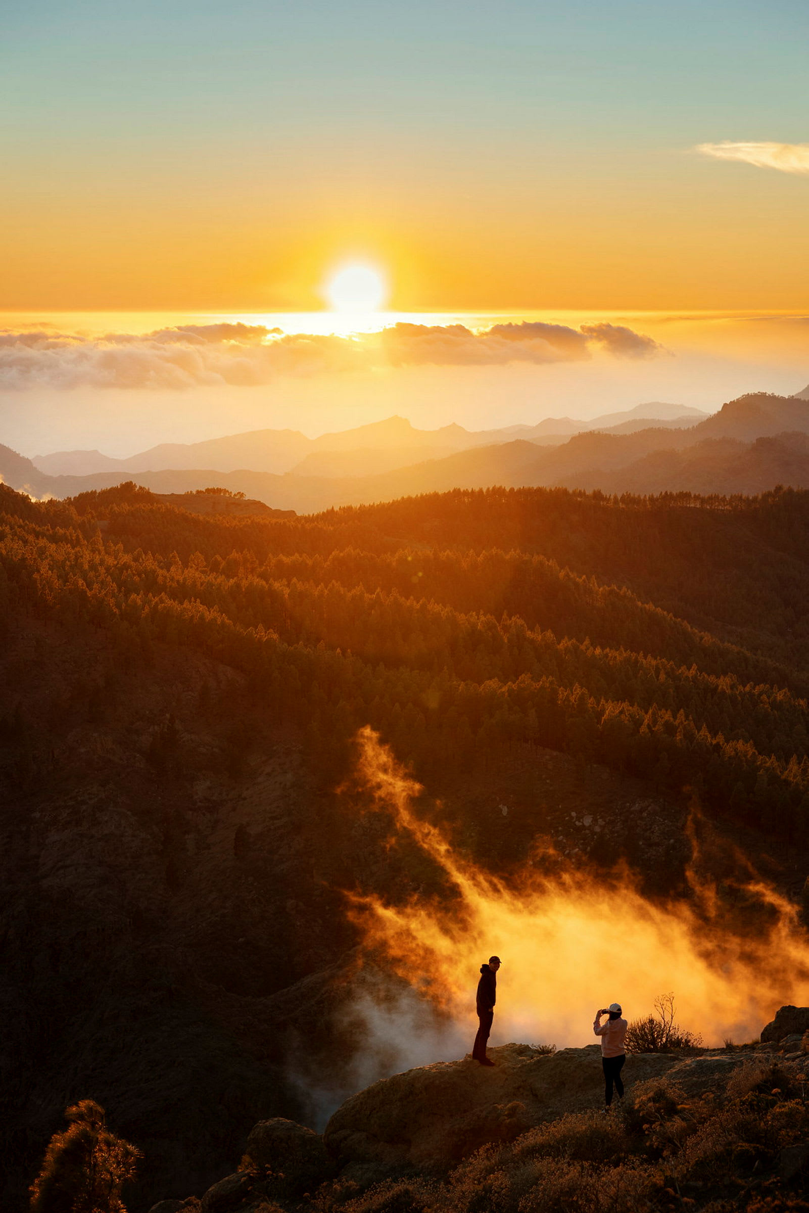 Solnedgang over fjell og skoger, med to personer som står på en høyde. (AI-generert tekst)