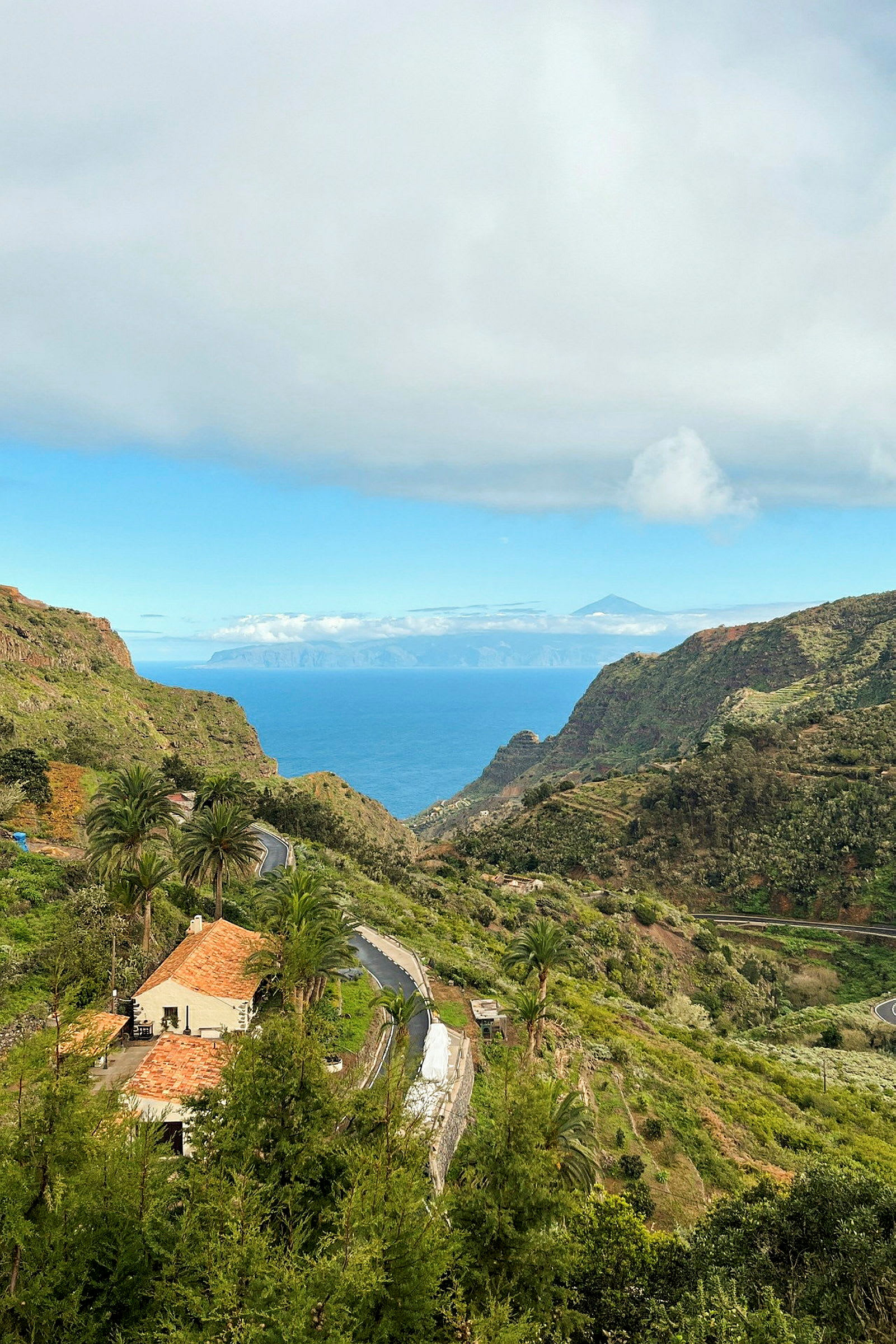 Landskap med grønne åser, et hus med rødt tak og en vei som slynger seg gjennom dalen, med havet og en fjelltopp i bakgrunnen. (AI-generert tekst)