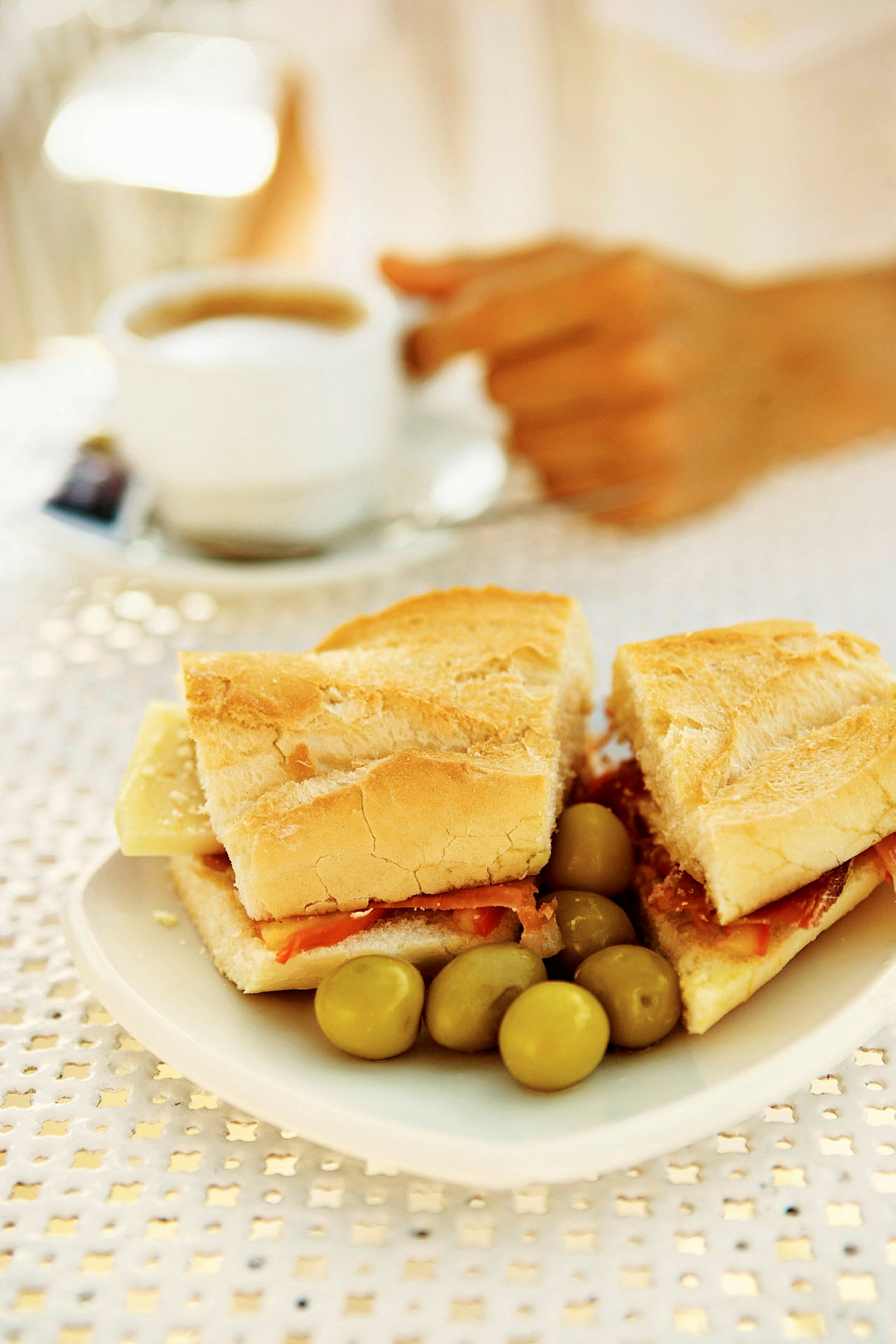 En tallerken med en baguette fylt med ost og skinke samt noen oliven. I bakgrunnen sees en hånd som holder en kopp kaffe. (AI-generert tekst)