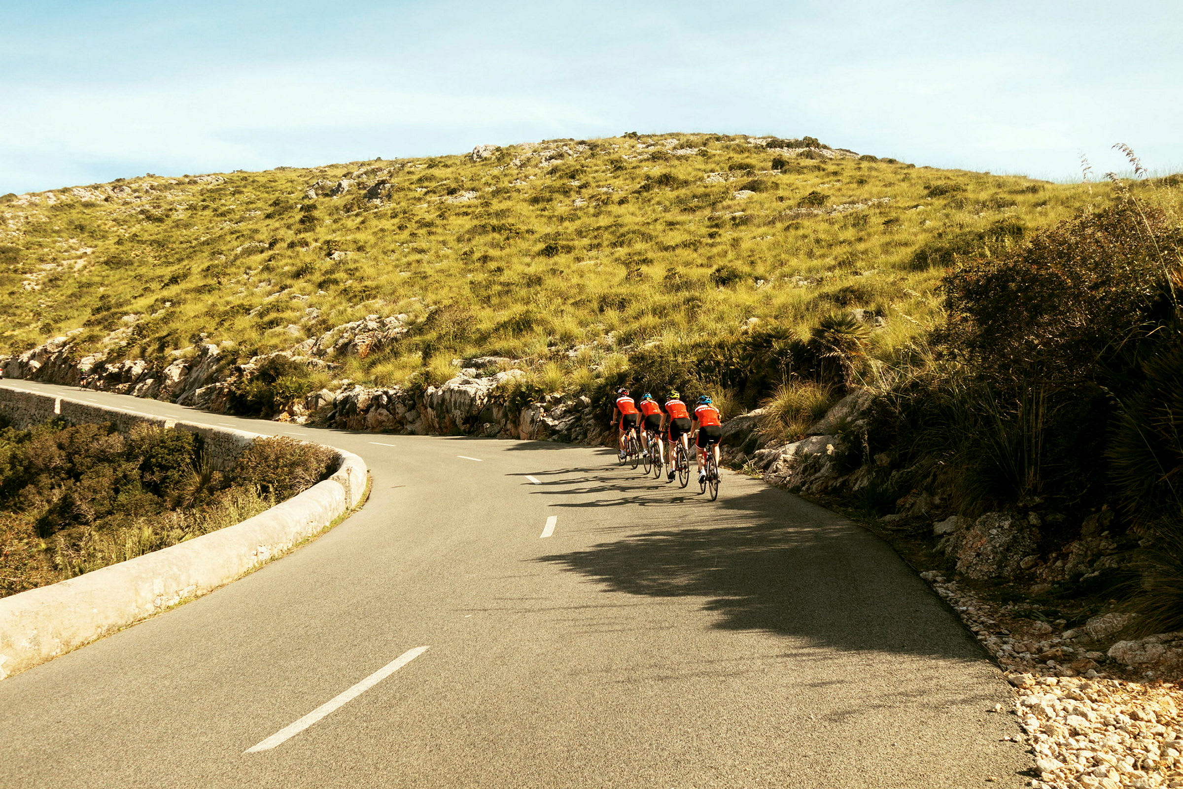 Sykkelgruppe på asfaltvei gjennom landskap med grønn vegetasjon og åser. (AI-generert tekst)