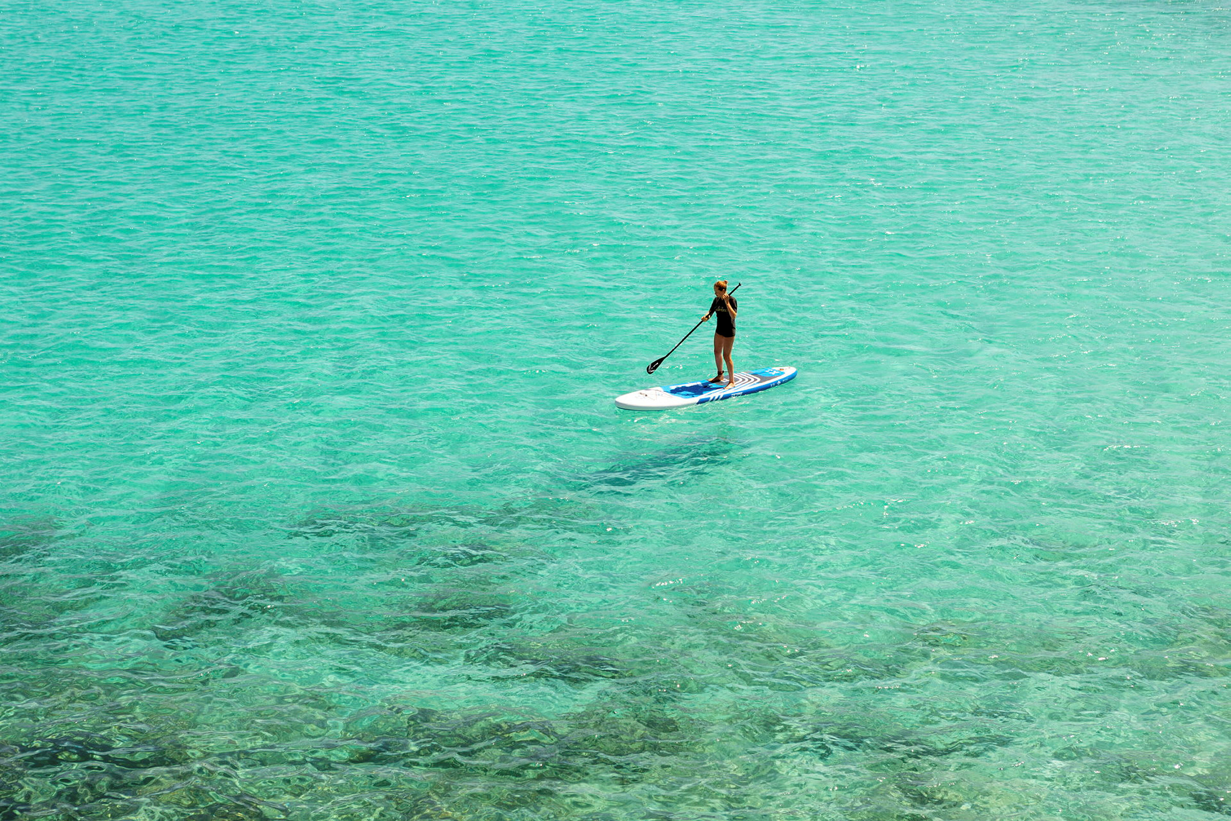 Person som padler stående på en paddleboard i klart turkis vann. (AI-generert tekst)