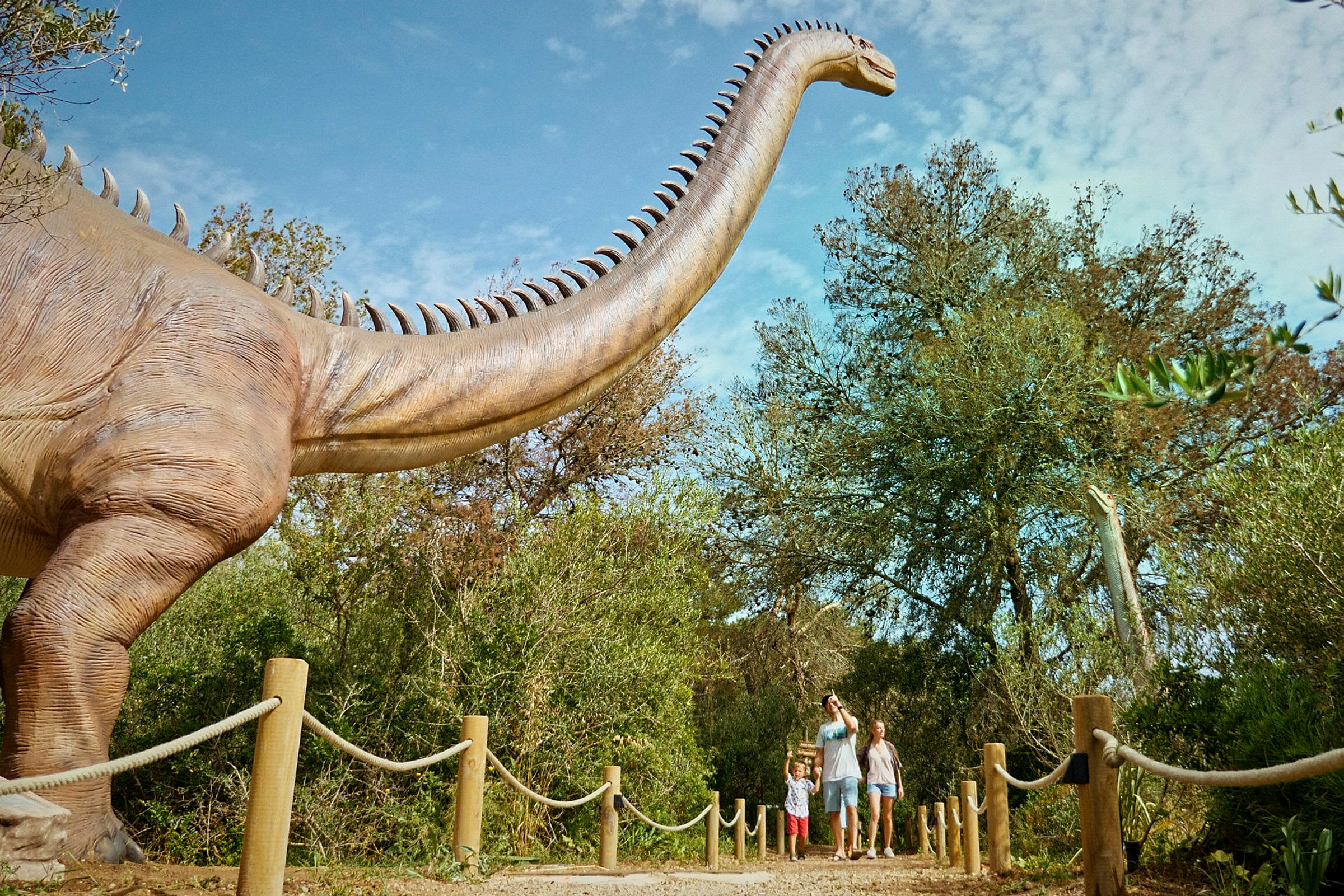 En familie spaserer på en sti ved siden av en stor dinosaurattrapp i en utendørsutstilling. (AI-generert tekst)