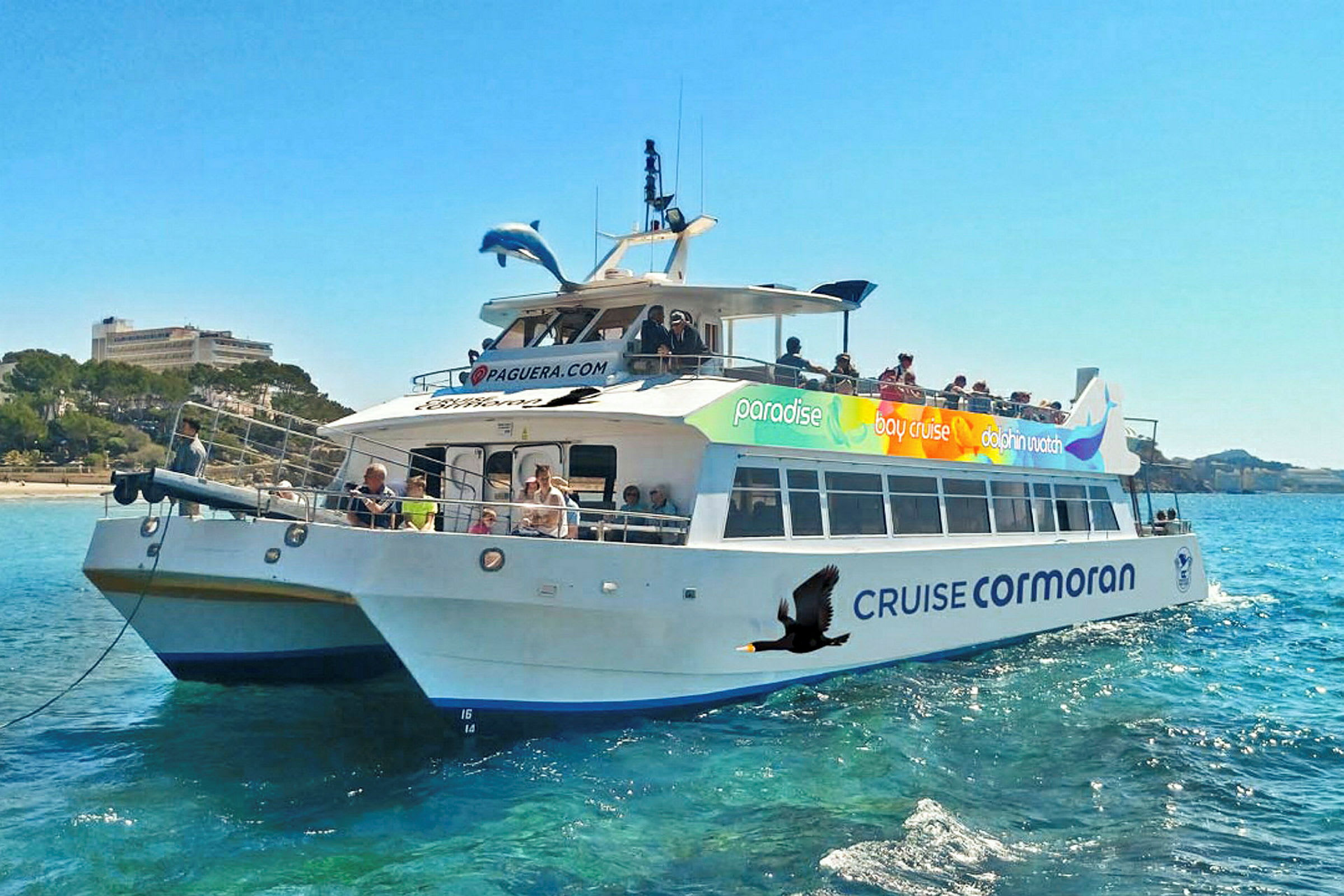 Et fartøy med navnet 'Cruise Cormoran' på vannet, omgitt av passasjerer og med blå himmel og landskap i bakgrunnen. (AI-generert tekst)