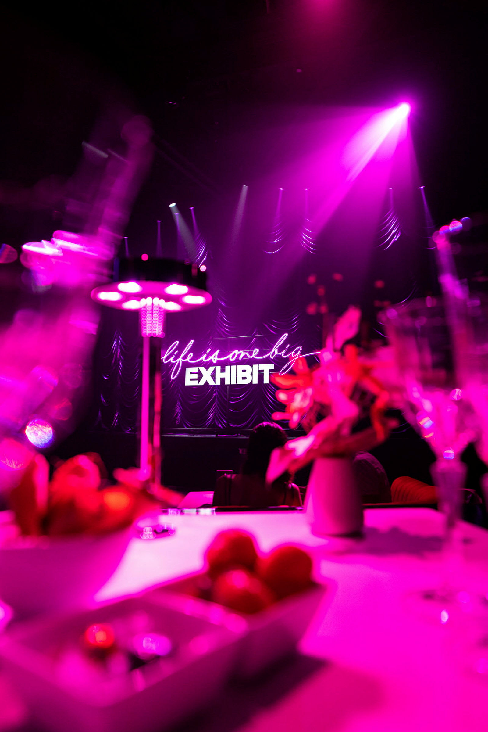 En opplyst scene med neonskiltet "life is one big exhibit" i bakgrunnen. Forgrunnen viser et bord med dekorasjoner og skåler. (AI-generert tekst)
