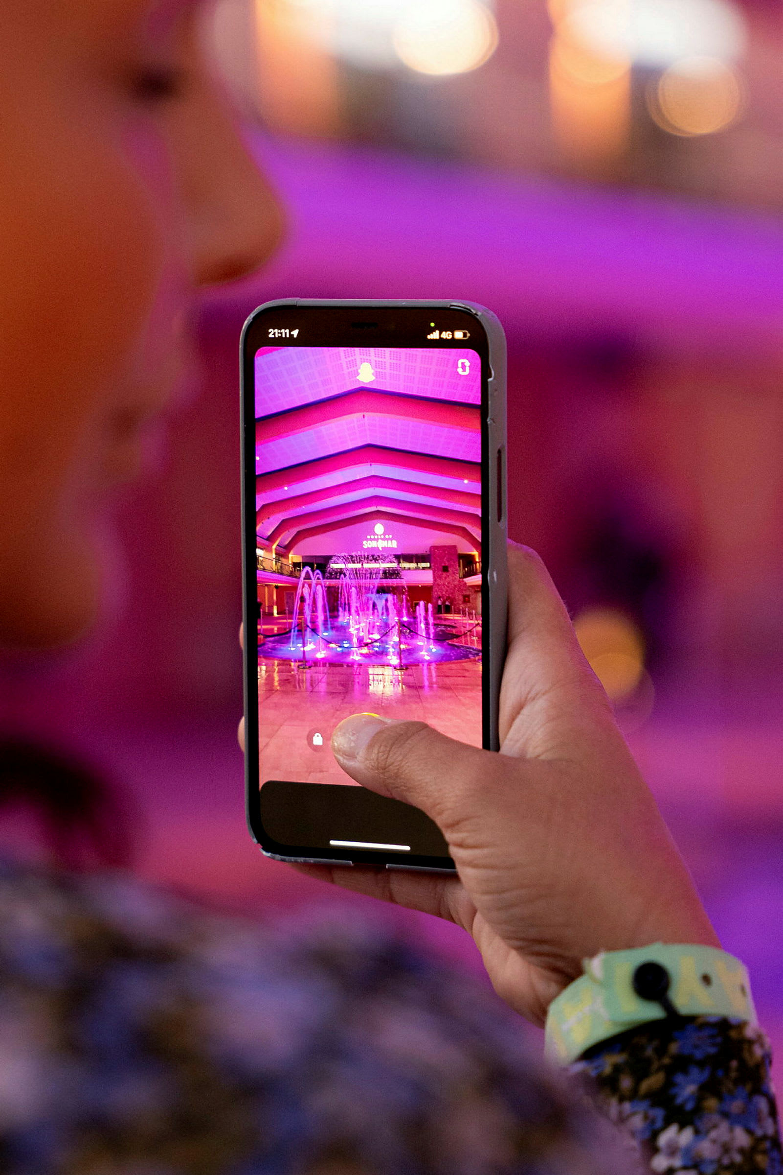 En person holder en mobiltelefon med et bilde av en opplyst fontene og en arkitektonisk takdesign på skjermen. (AI-generert tekst)