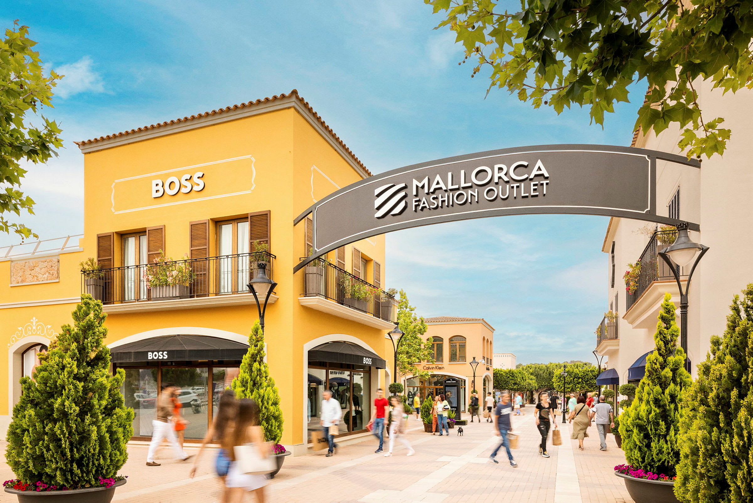 En handlegate med gule bygninger som viser skilt for butikker, blant annet BOSS. Et skilt med teksten "Mallorca Fashion Outlet" sees over inngangen til området. (AI-generert tekst)