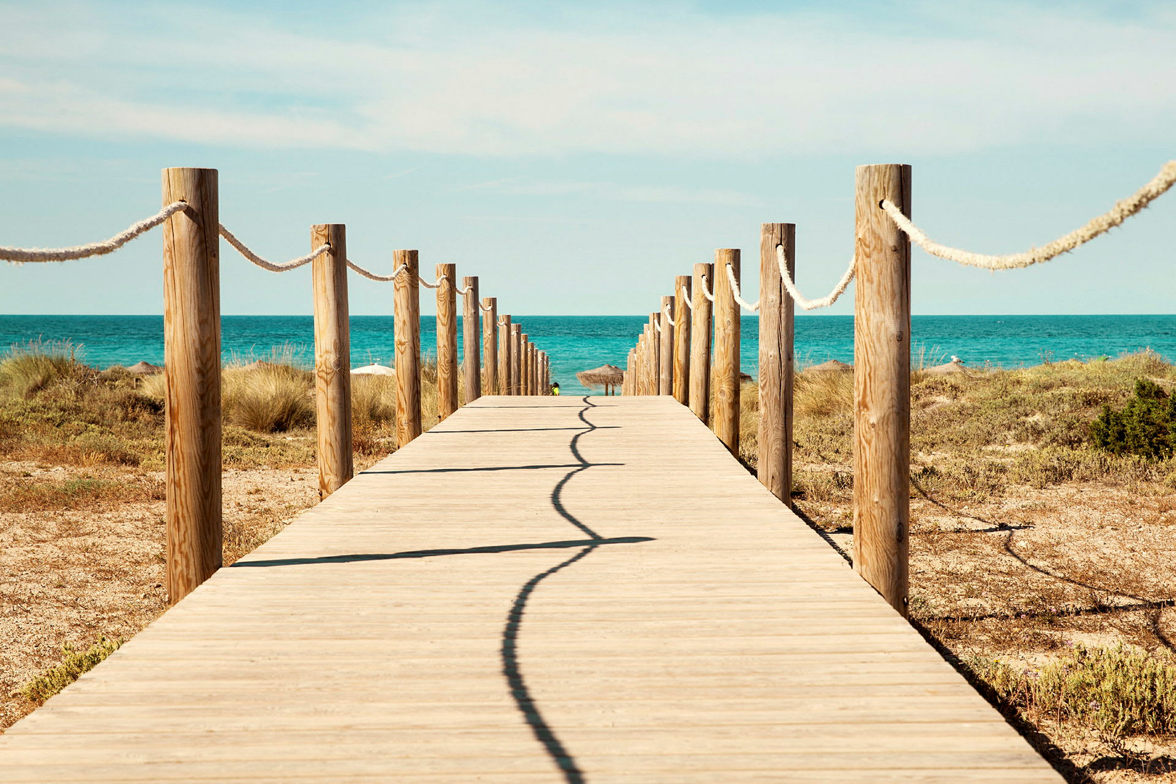 En trebro fører mot havet, kantet av trestolper og tau, med sand og vegetasjon på sidene. (AI-generert tekst)