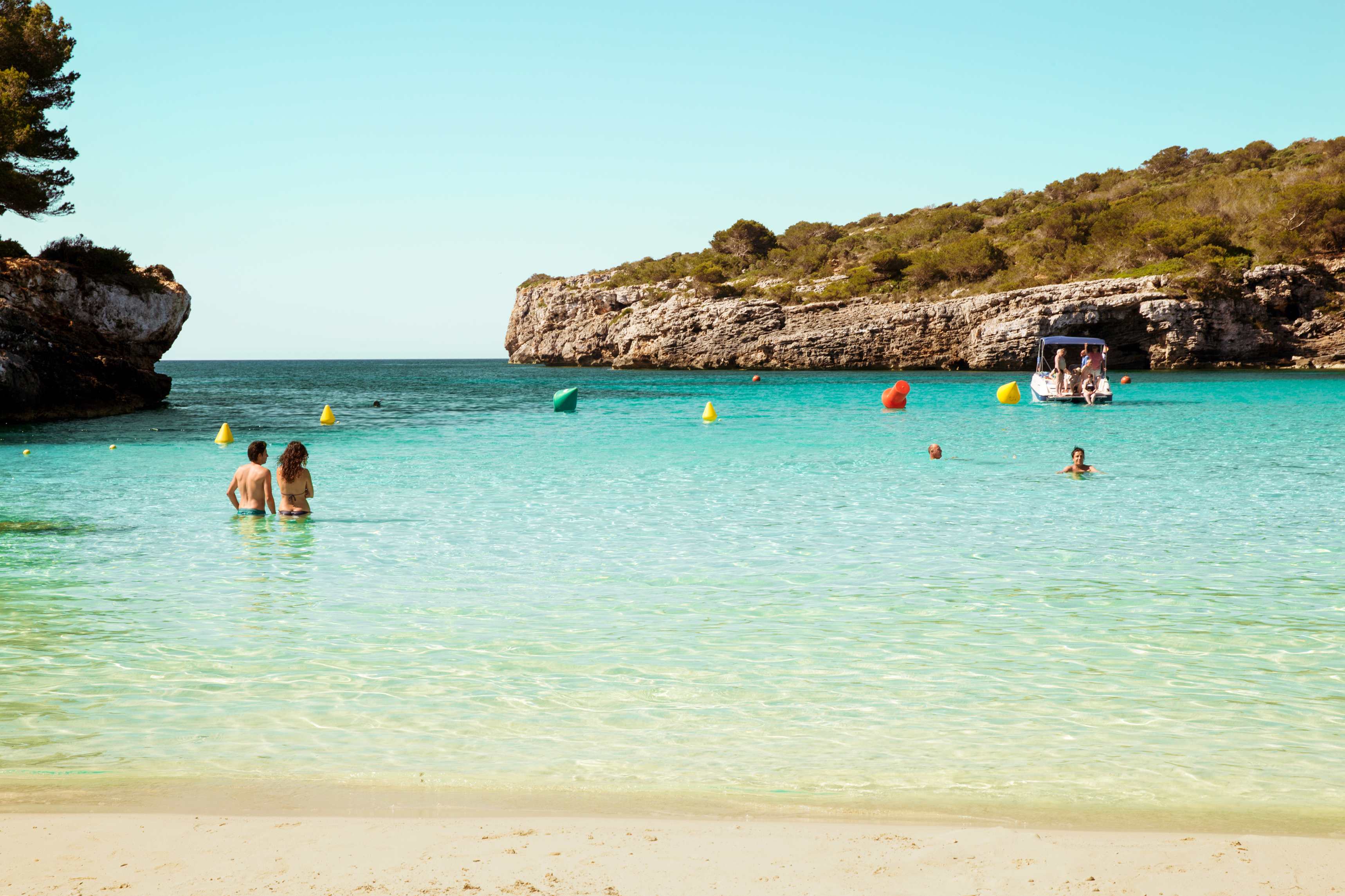 En sandstrand med mennesker i det rolige, turkise vannet. I bakgrunnen er en båt og klipper dekket av grønt. (AI-generert tekst)