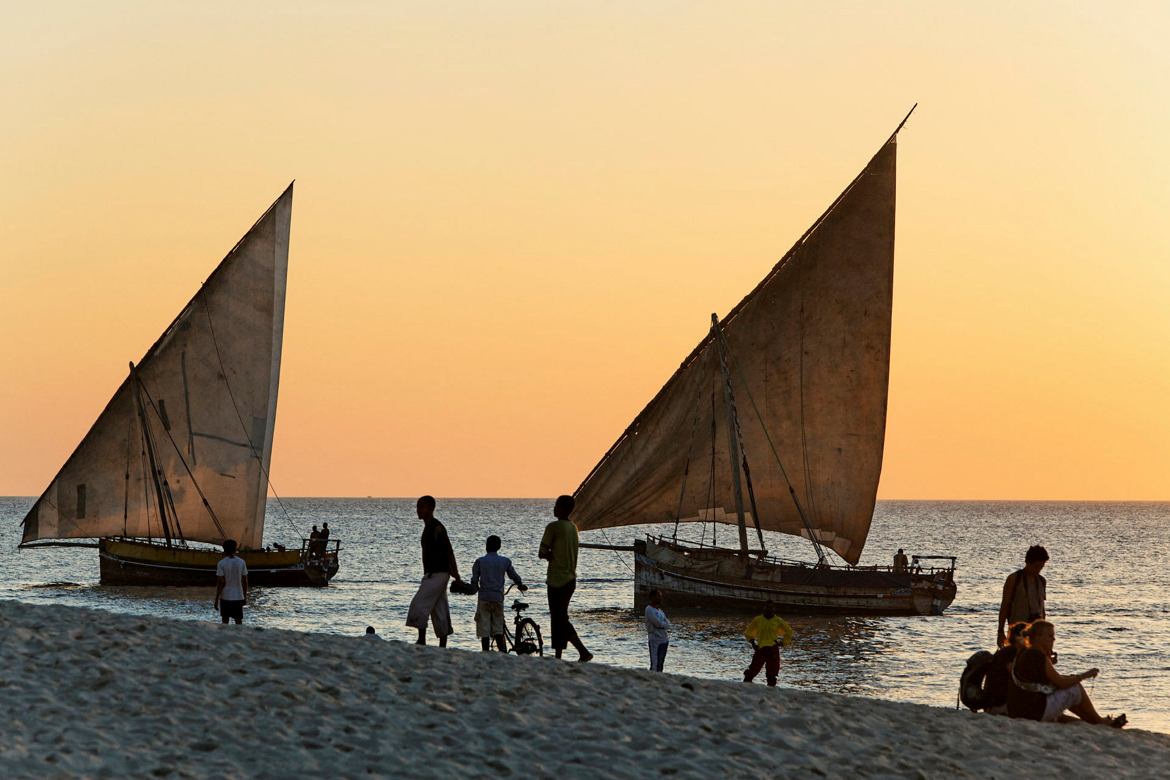 Folk går på en strand ved solnedgang. To trebåter med seil ligger ved vannet. (AI-generert tekst)