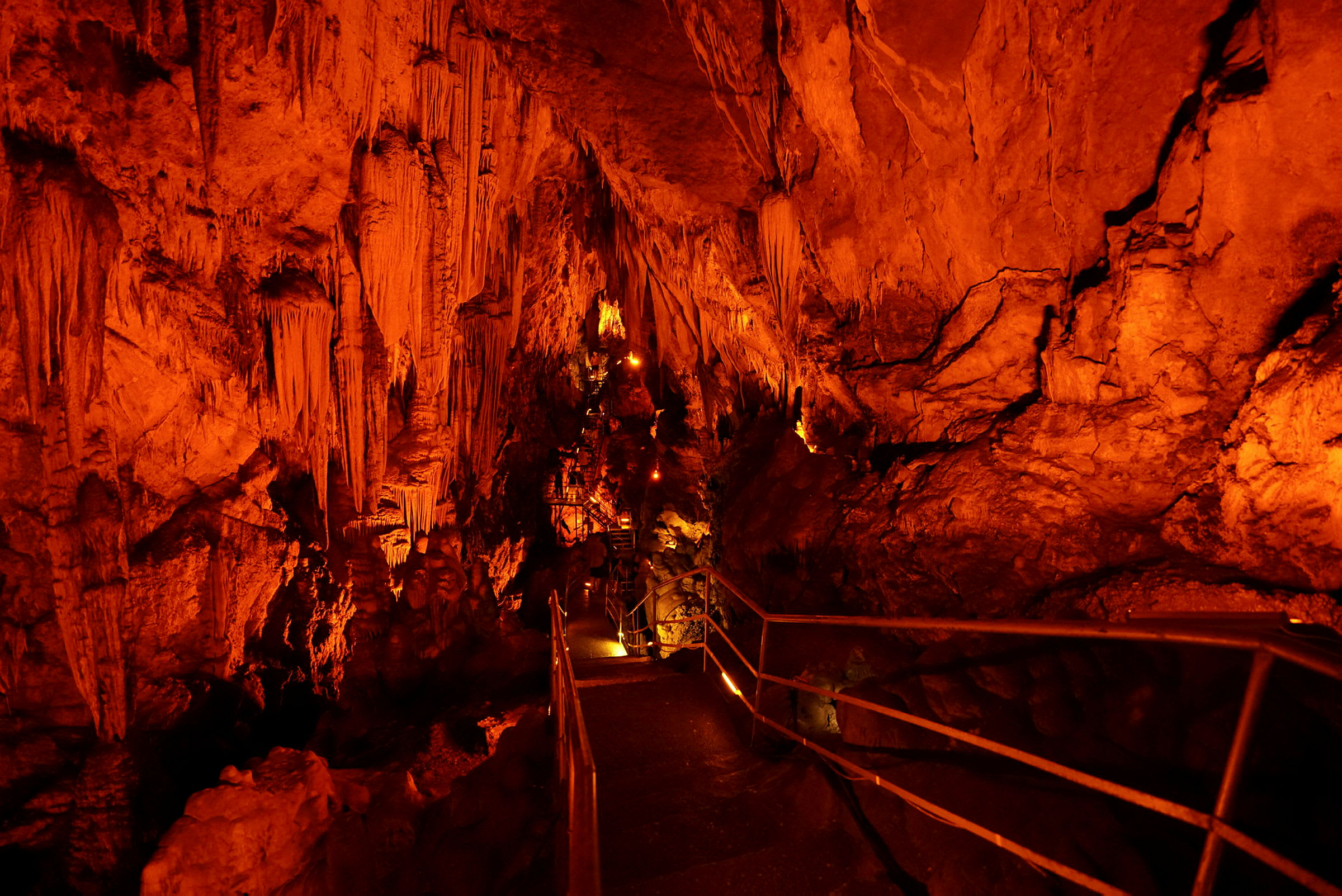 En hule med stalaktitter og stalagmitter, opplyst med oransje belysning. En gangsti med rekkverk fører gjennom hulen. (AI-generert tekst)