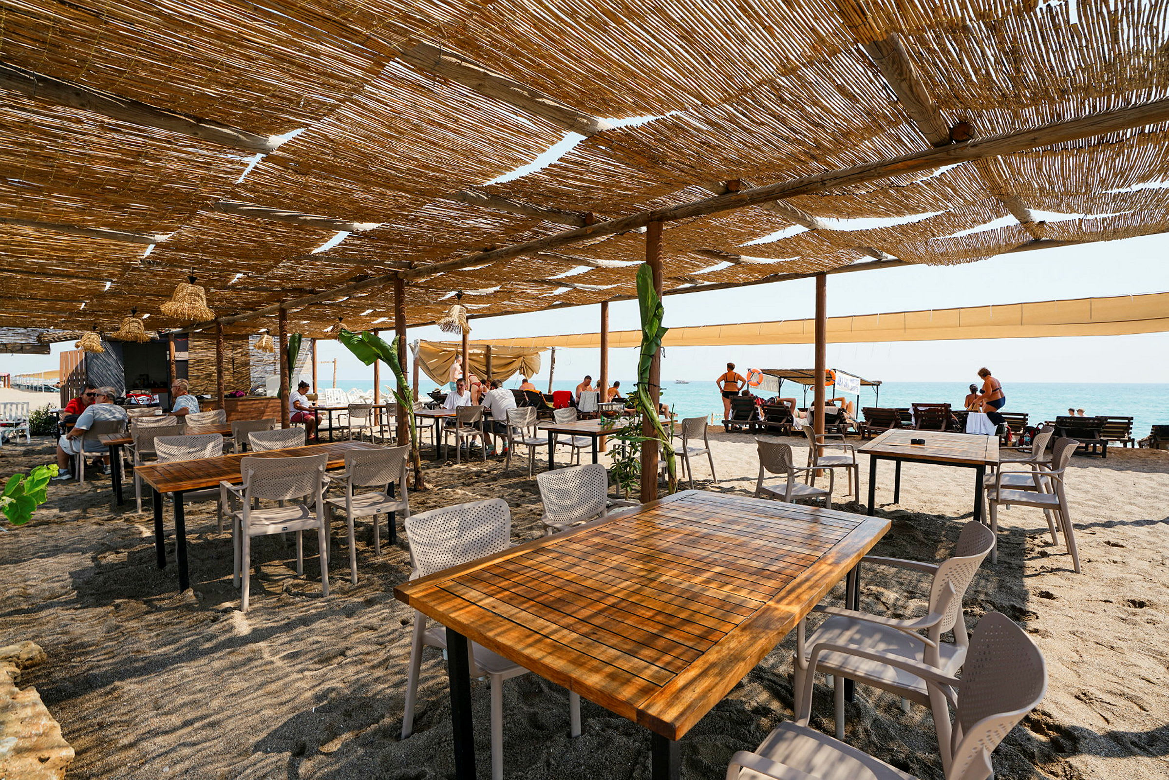 Utendørs restaurant med bord og stoler på sandstrand under tak av bambus. (AI-generert tekst)