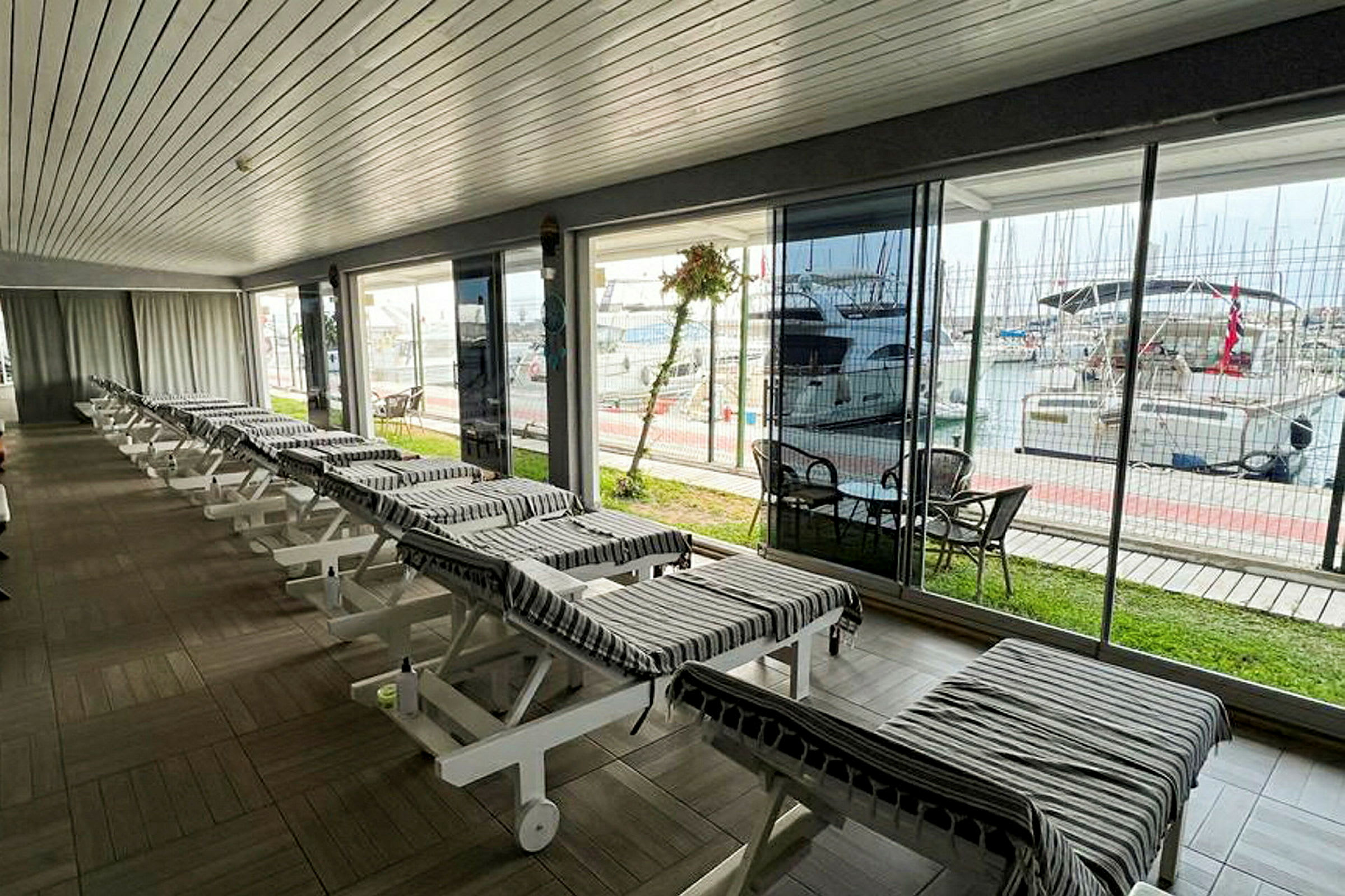Et lyst innemiljø med solsenger arrangert i rekker, og en marina med båter vises utenfor. (AI-generert tekst)
