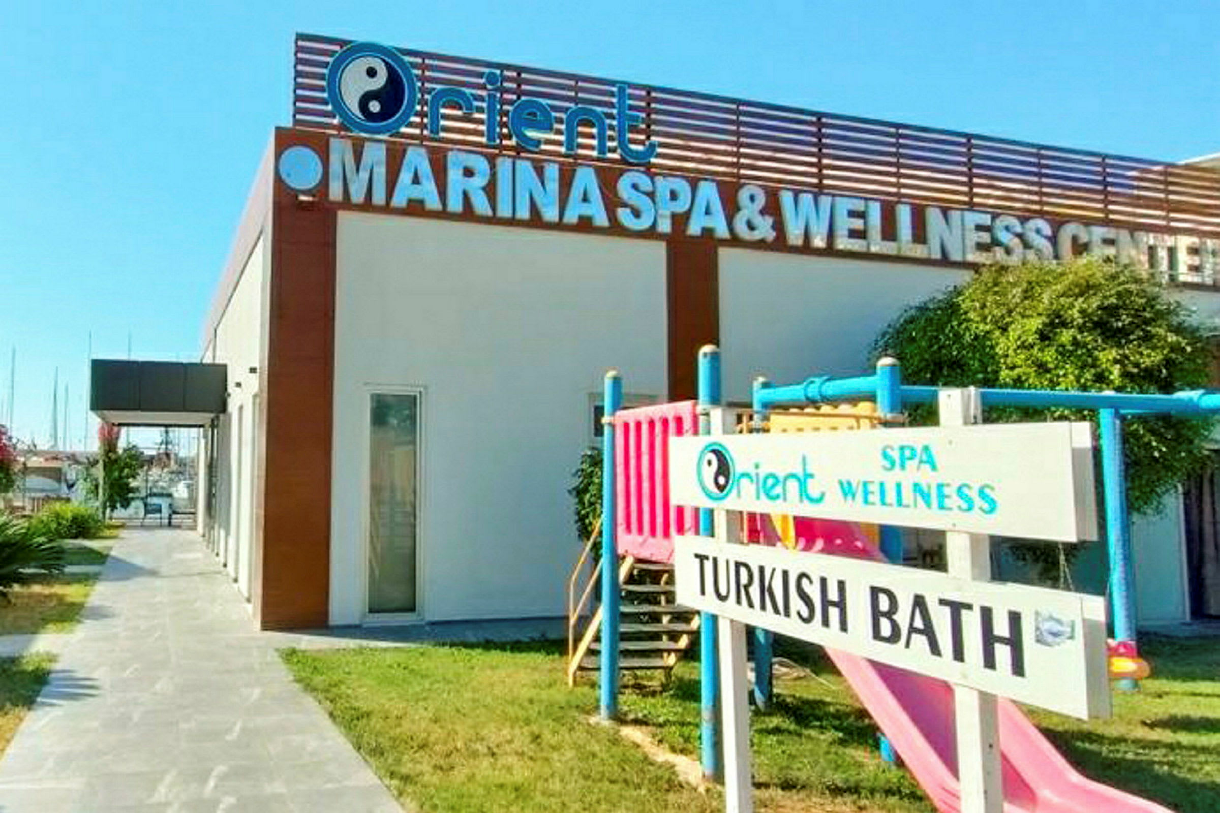 Bygning med et skilt som sier 'MARINA SPA & WELLNESS CENTER'. På plenen er det også en lekeplass for barn og et skilt med teksten 'TURKISH BATH'. (AI-generert tekst)