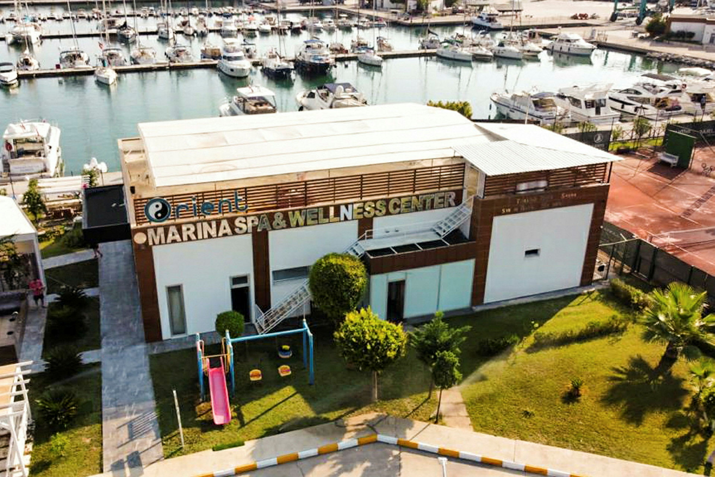 Bygning med skiltet 'Marina Spa & Wellness Center', lekeplass med sklie og bakgrunn med småbåthavn. (AI-generert tekst)