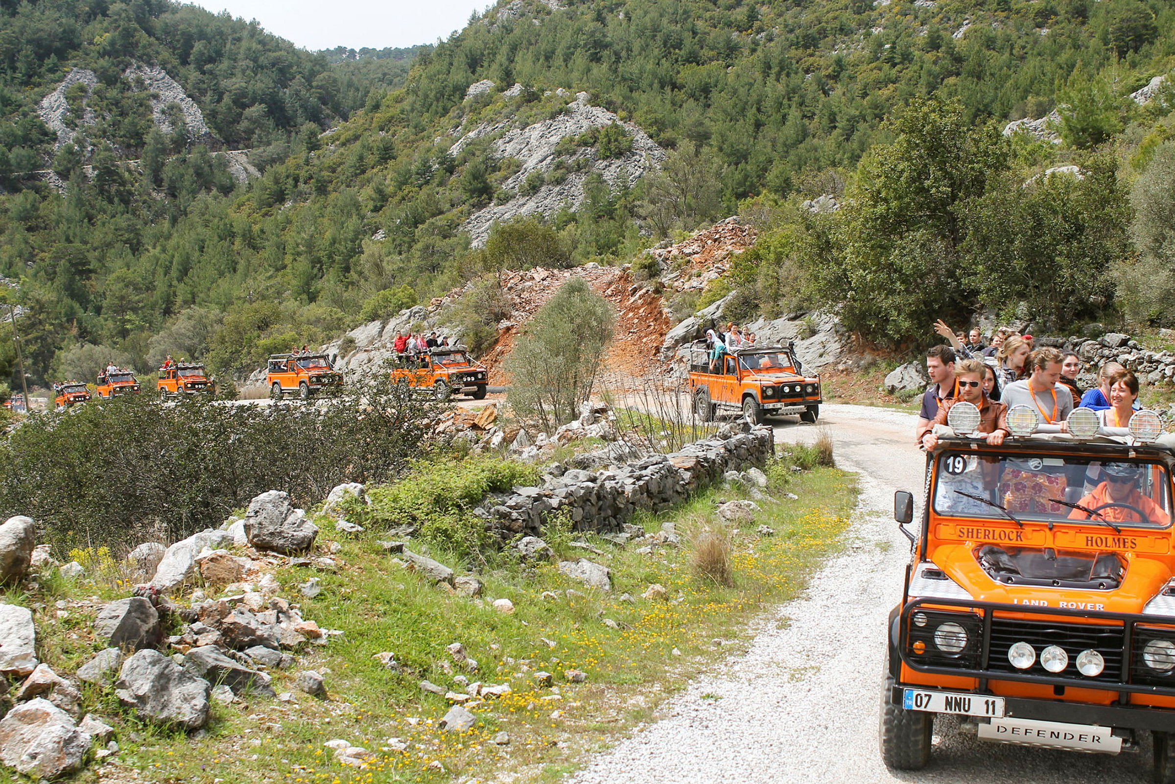 En gruppe oransje jeep kjører på en grusvei gjennom et fjellrikt og frodig landskap. (AI-generert tekst)