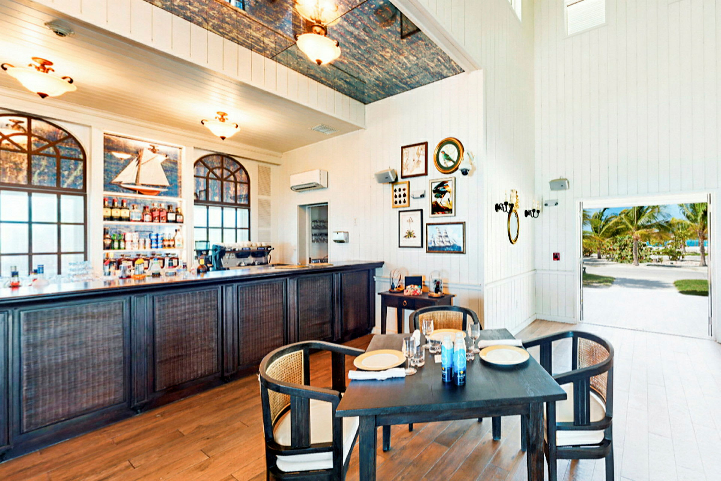 Et innendørsområde med en bar, spisebord, dekorasjoner på veggen og en åpning som viser utendørsmiljøet. (AI-generert tekst)